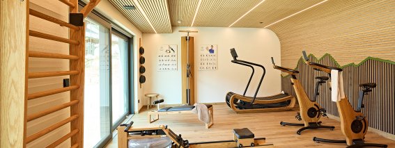 In einem Raum gibt es Fitnessgeräte wie Laufband, Rudergerät und Fahrräder. Das Licht ist hell und der Boden ist aus Holz.
