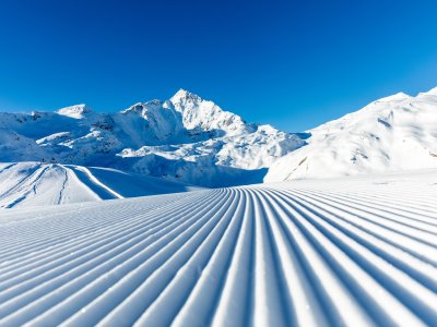 Am Morgen zeigt sich eine friedliche Winterlandschaft mit schneebedeckten Bergen und präparierten Pisten unter strahlend blauem Himmel.