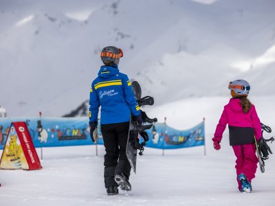 Auf der Piste üben zwei Kinder das Snowboard fahren. Sie tragen Helme und sind bereit für den nächsten Versuch.