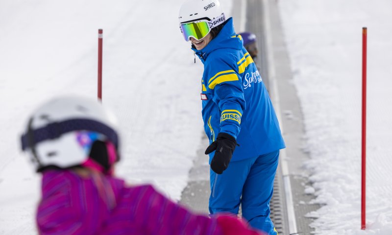 Ein Skilehrer zeigt Kindern die Grundlagen des Skifahrens auf einer verschneiten Piste an einem sonnigen Tag.