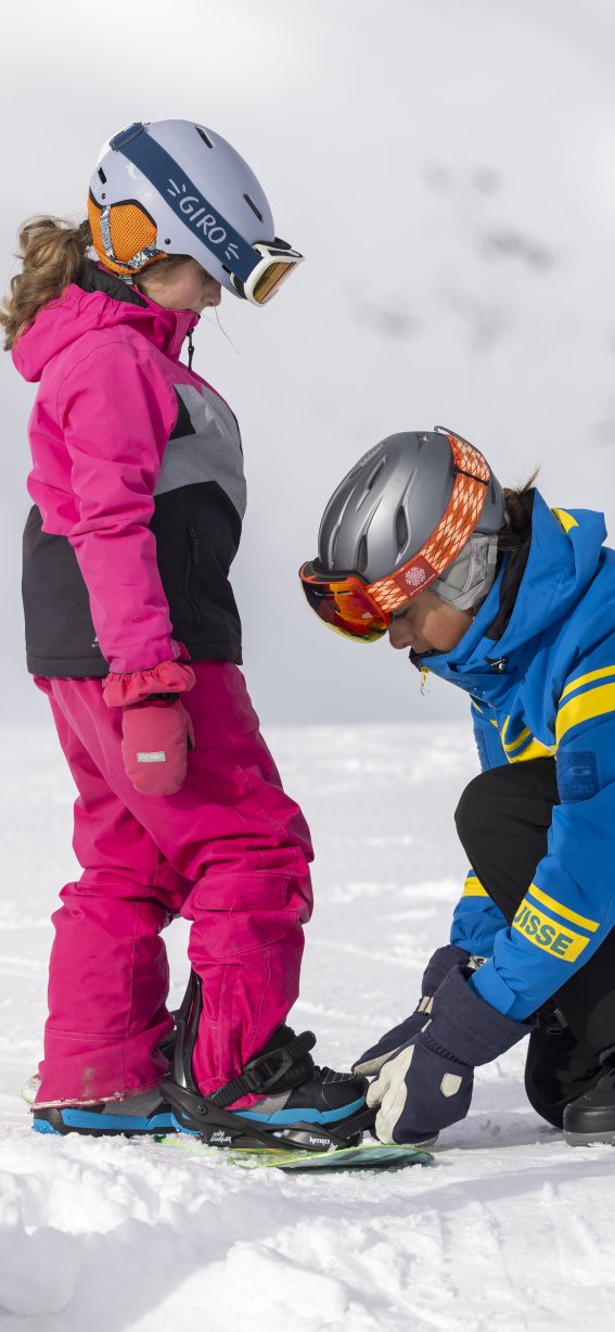 Ein Erwachsener hilft einem Kind, sich für das Snowboarden vorzubereiten. Es ist sonnig und verschneit in den Bergen.