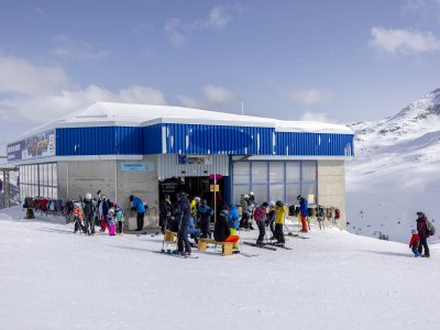 Viele Skifahrer stehen vor einer Bergstation und genießen den sonnigen Tag in den Alpen. Die Umgebung ist schneebedeckt.