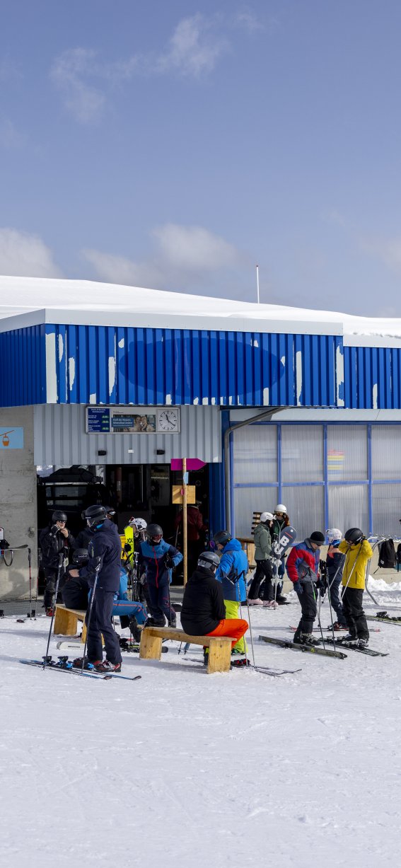 Viele Skifahrer stehen vor einer Bergstation und genießen den sonnigen Tag in den Alpen. Die Umgebung ist schneebedeckt.