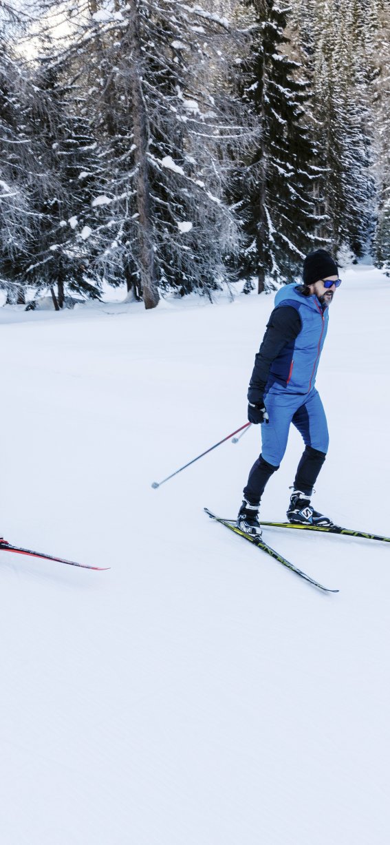 Zwei Personen skifahren im verschneiten Wald. Die Sonne scheint auf die weißen Pisten und die Bäume.