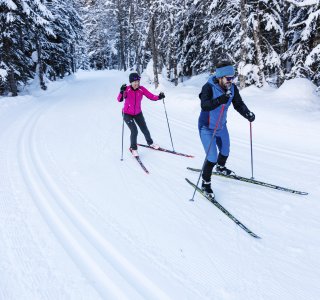 Zwei Personen ski wandern auf einer tief verschneiten Strecke im Wald. Die Szene zeigt die Ruhe und Schönheit der Winterlandschaft.