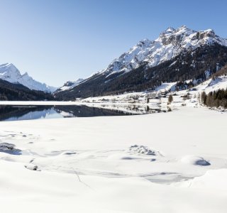 Die verschneite Alpenlandschaft zeigt einen ruhigen See und hohe Berge unter klarem blauen Himmel im Winter.