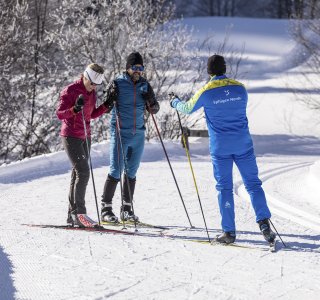 Zwei Personen lernen beim Skilanglauf von einem Lehrer in einer winterlichen Kulisse mit Schnee und Bäumen.