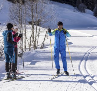 Drei Langläufer stehen auf einer präparierten Loipe in der Winterlandschaft. Die Sonne scheint auf den Schnee.