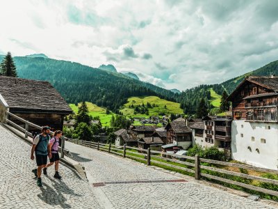 Zwei Wanderer gehen durch ein idyllisches Bergdorf mit grünen Hügeln und Wolken am Himmel an einem Sommertag.