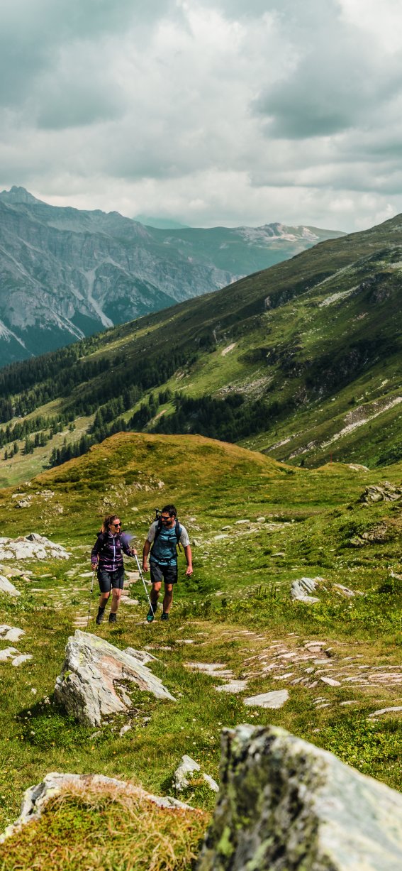 Zwei Wanderer gehen auf einem Pfad durch die grünen Berge an einem bewölkten Tag.