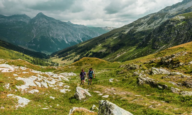 Zwei Wanderer gehen auf einem Pfad durch die grünen Berge an einem bewölkten Tag.