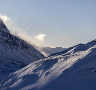 Eine verschneite Berglandschaft mit weichen Hügeln und wolkenverhangenen Gipfeln bei Sonnenaufgang im Winter.