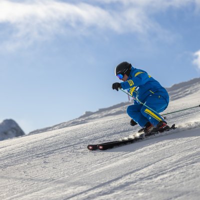 Ein Skifahrer in türkisfarbener Kleidung fährt einen Hang hinunter. Die Sonne scheint und die Berge sind im Hintergrund sichtbar.