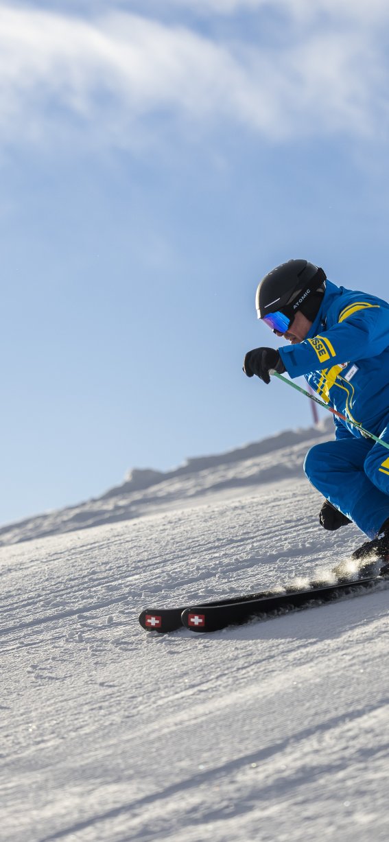 Ein Skifahrer in türkisfarbener Kleidung fährt einen Hang hinunter. Die Sonne scheint und die Berge sind im Hintergrund sichtbar.