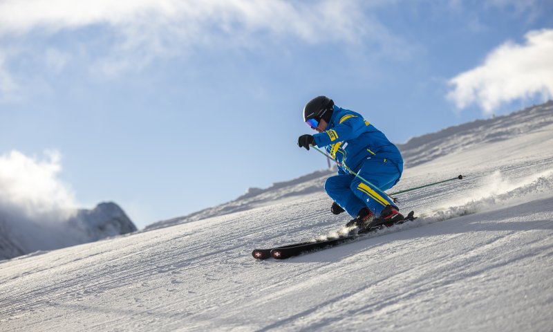 Ein Skifahrer in türkisfarbener Kleidung fährt einen Hang hinunter. Die Sonne scheint und die Berge sind im Hintergrund sichtbar.
