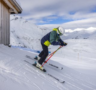 Ein Skifahrer beginnt die Abfahrt von einer Berghütte in den Alpen bei klarem Himmel und schneebedeckten Bergen.