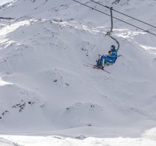 Ein Skifahrer fährt mit dem Sessellift durch verschneite Alpenlandschaft im Winter. Die Berge sind hoch und weiß bedeckt.
