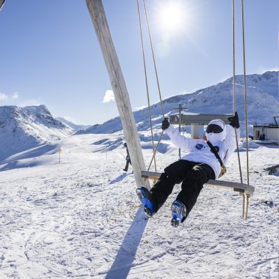Eine Person schwingt entspannt auf einer Schaukel im Schnee. Die Berge sind im Hintergrund sichtbar, der Himmel ist klar.