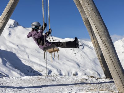 Ein Kind schwingt fröhlich auf einer Schaukel im Schnee vor einer Berglandschaft. Es ist ein klarer Wintertag.