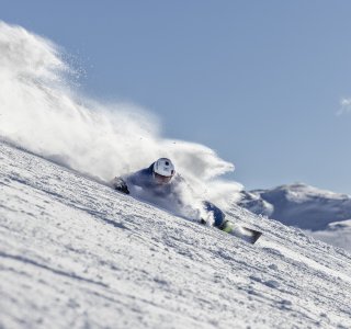Ein Schneebrettfahrer fährt schnell den schneebedeckten Hang hinunter. Die Sonne scheint und der Schnee spritzt auf.