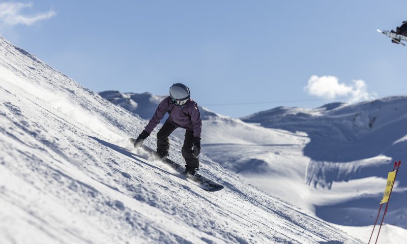 Ein Snowboardfahrer fährt auf einer schneebedeckten Piste in den Alpen bei sonnigem Wetter und klarer Sicht.