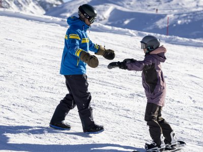Zwei Kinder üben am Hang das Snowboardfahren unter blauen Himmel und sonnigem Wetter in den Bergen.