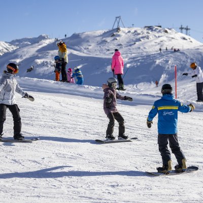 Kinder und Erwachsene lernen beim Snowboarden auf der verschneiten Piste in den Alpen an einem sonnigen Wintertag.