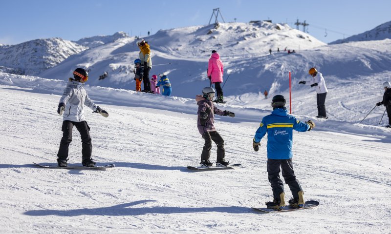 Kinder und Erwachsene lernen beim Snowboarden auf der verschneiten Piste in den Alpen an einem sonnigen Wintertag.