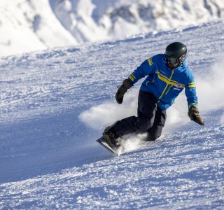 Ein Snowboardfahrer fährt die verschneite Piste hinunter und hinterlässt eine Wolke aus Schnee. Berglandschaft im Hintergrund.