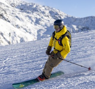 Ein Skifahrer fährt mit Schwung über die schneebedeckten Pisten in den Bergen an einem sonnigen Tag.
