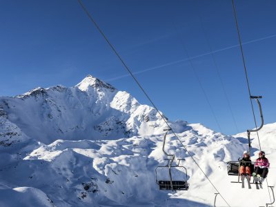 Zwei Skifahrer fahren mit dem Sessellift in den verschneiten Alpen auf einem sonnigen Tag.