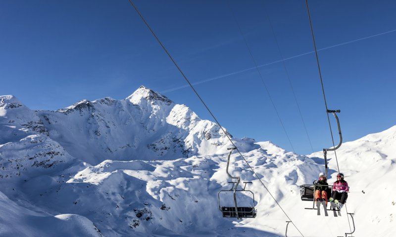 Zwei Skifahrer fahren mit dem Sessellift in den verschneiten Alpen auf einem sonnigen Tag.
