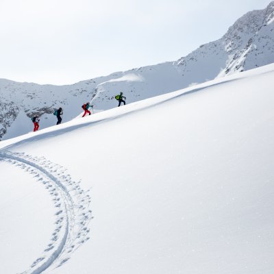 Drei Personen wandern durch frischen Schnee in den Bergen. Die Sonne scheint und die Landschaft ist klar und winterlich.