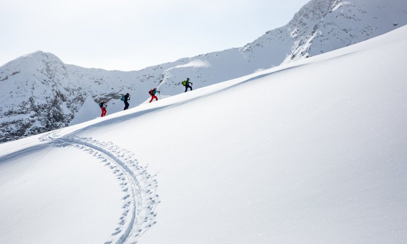 Drei Personen wandern durch frischen Schnee in den Bergen. Die Sonne scheint und die Landschaft ist klar und winterlich.
