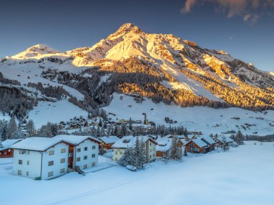 Eine verschneite Berglandschaft mit Häusern im Vordergrund und der Sonne, die auf die Gipfel scheint, schafft eine ruhige Atmosphäre.