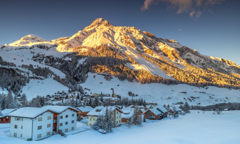 Eine verschneite Berglandschaft mit Häusern im Vordergrund und der Sonne, die auf die Gipfel scheint, schafft eine ruhige Atmosphäre.