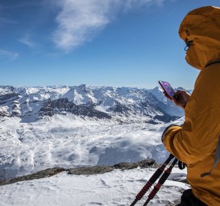 Ein Wanderer trägt eine orange Jacke und schaut auf sein Handy, während er die bergige Winterlandschaft bewundert.