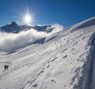 Menschen wandern auf einer schneebedeckten Fläche in den Bergen. Die Sonne scheint am klaren Himmel.