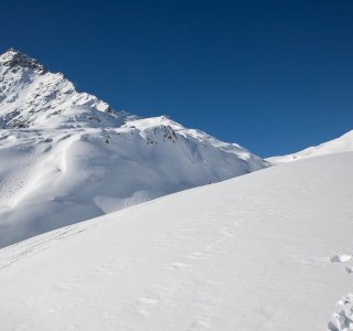 Zwei Skifahrer wandern durch frischen Schnee in den Alpen. Der Himmel ist blau und die Berge sind schneebedeckt.