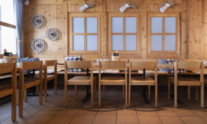 Der Innenraum zeigt Holzstühle und Tische in einem Restaurant. Fenster sind an der Wand. Es ist tagsüber.