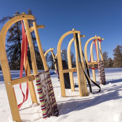 Schlitten stehen im Schnee in den Bergen. Die Sonne scheint auf die Holzkonstruktionen. Der Himmel ist blau.