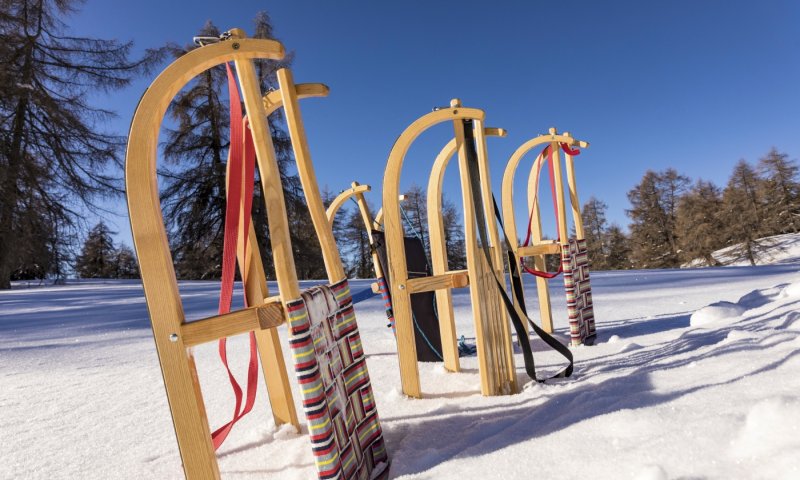 Schlitten stehen im Schnee in den Bergen. Die Sonne scheint auf die Holzkonstruktionen. Der Himmel ist blau.