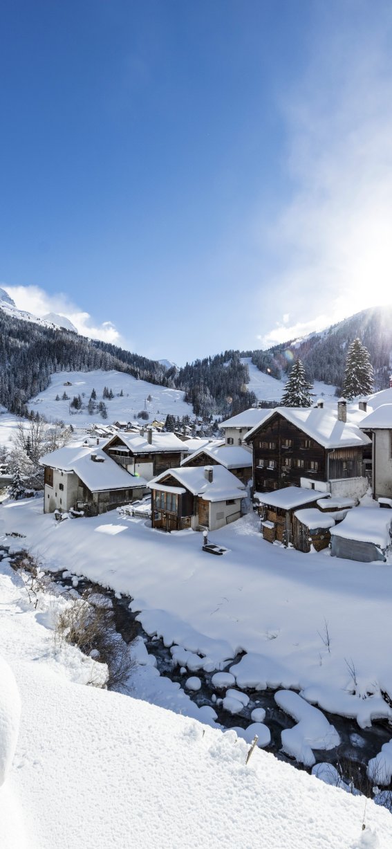 In einem Dorf liegt Schnee auf den Dächern. Es gibt Berge im Hintergrund und eine ruhige Straße.
