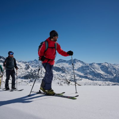 Drei Personen fahren auf Skiern in den Bergen. Der Himmel ist blau, und die Landschaft ist schneebedeckt.