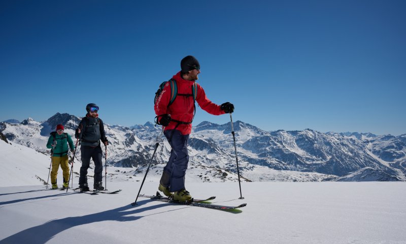 Drei Personen fahren auf Skiern in den Bergen. Der Himmel ist blau, und die Landschaft ist schneebedeckt.