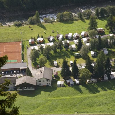 Auf dem Campingplatz stehen viele Wohnwagen und Zelte. Der Platz liegt in einem Tal mit Bergen und einem Fluss in der Nähe.