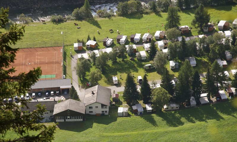 Auf dem Campingplatz stehen viele Wohnwagen und Zelte. Der Platz liegt in einem Tal mit Bergen und einem Fluss in der Nähe.
