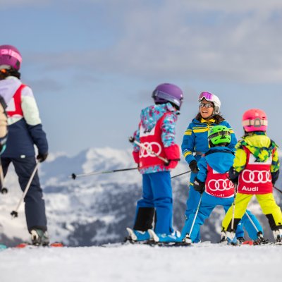 Skischulkinder tragen Startnummern und üben mit einer Skilehrerin auf der Piste.