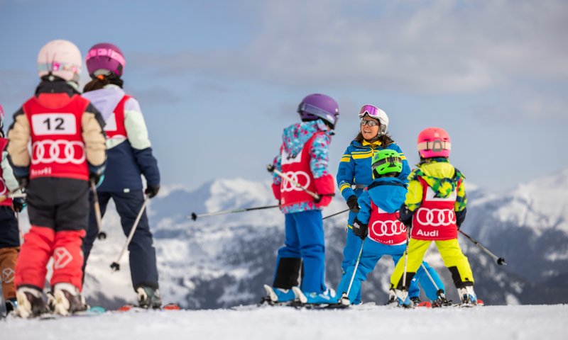 Skischulkinder tragen Startnummern und üben mit einer Skilehrerin auf der Piste.