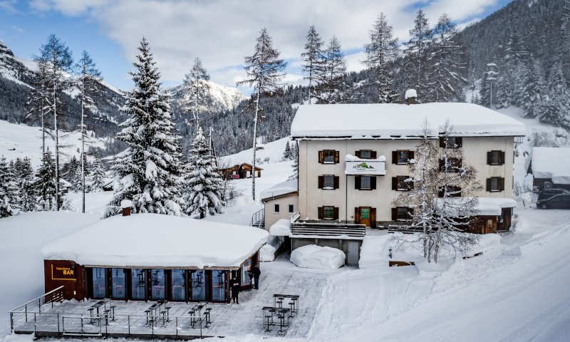 Das Bild zeigt das Gebäude Hüschera Lodge, das direkt an der Piste und neben der Hüschera Bard liegt. Die Landschaft darum ist tief verschneit.